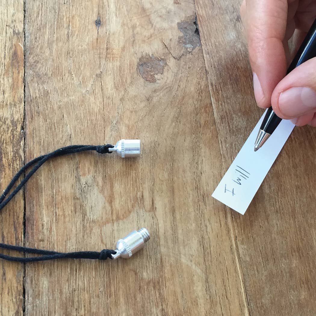 Black lanyard with metal ends on a wooden surface, next to a hand holding a pen and paper.