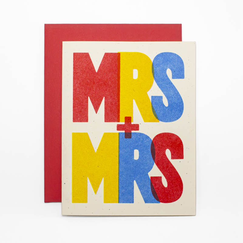 A square greeting card with a red envelope behind it, featuring the words “MRS + MRS” in large block letters colored red, yellow, and blue on a cream cardstock background.