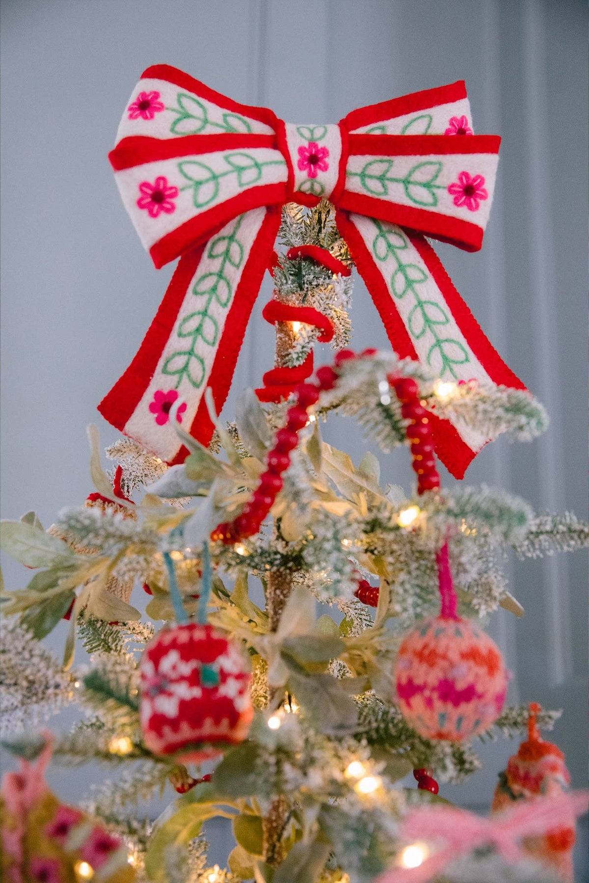 A close-up of a decorated Christmas tree with a large red and white bow topped with pink flowers and green leaf embroidery, alongside warm white string lights and knitted ornaments.