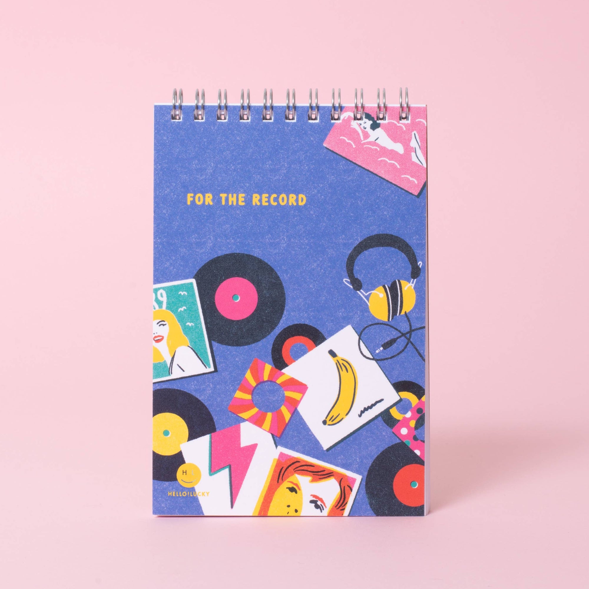 A spiral-bound notebook with a blue cover featuring retro vinyl records, headphones, and colorful illustrated cards, standing on a pink background.