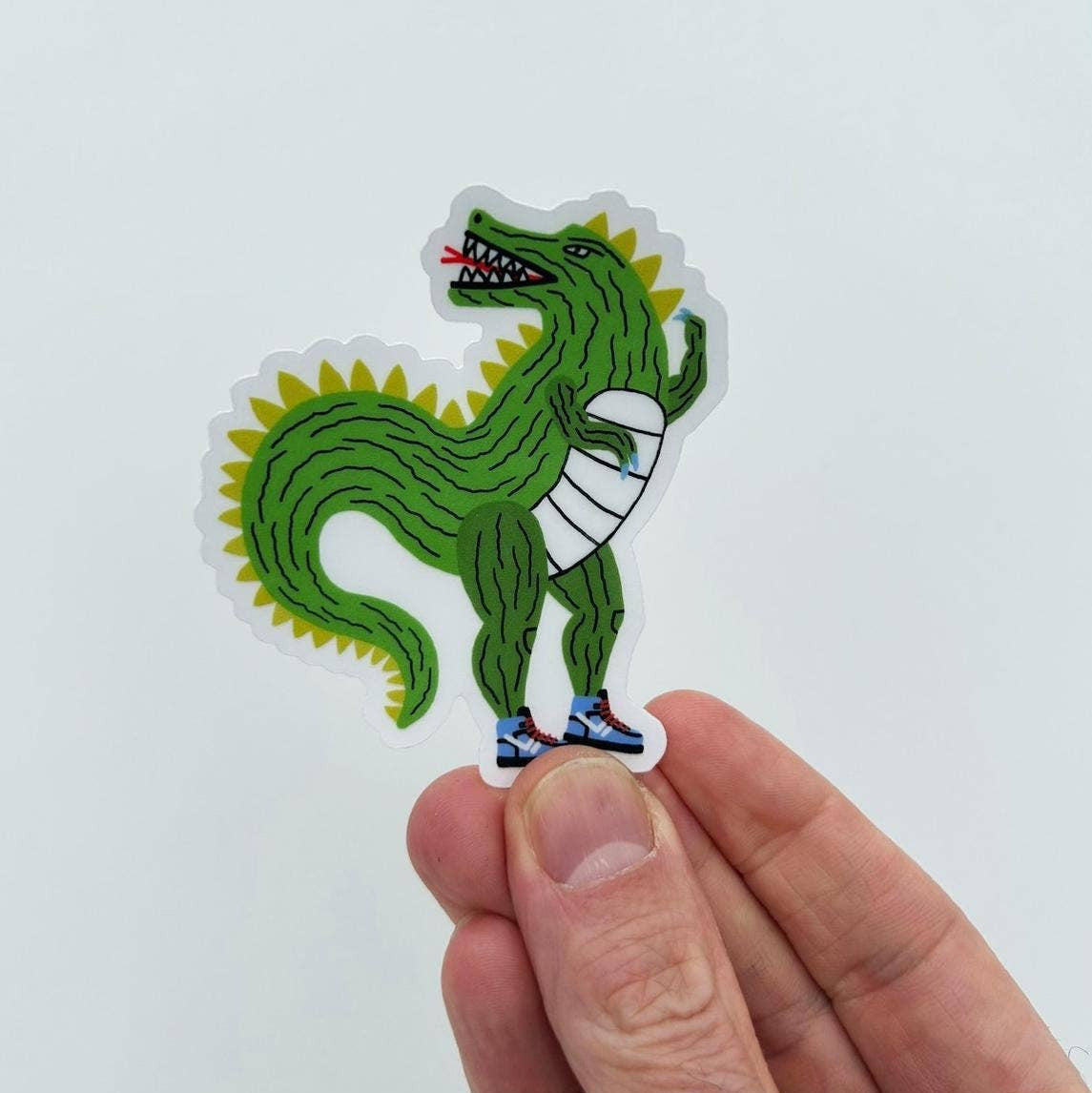 A die-cut sticker featuring a green cartoon dinosaur with yellow spikes, a white bandage on its arm, and blue sneakers, held between a person's fingers against a plain light background.