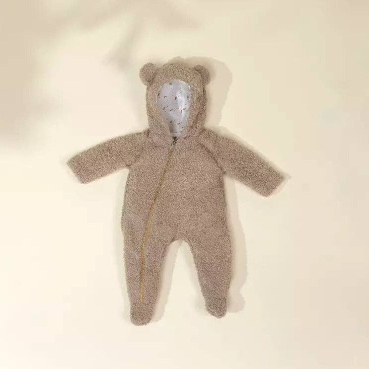A plush teddy bear hooded one-piece romper in a warm taupe-brown color, featuring a white embroidered face on the hood and a gold zipper, laid flat on a cream-colored background.
