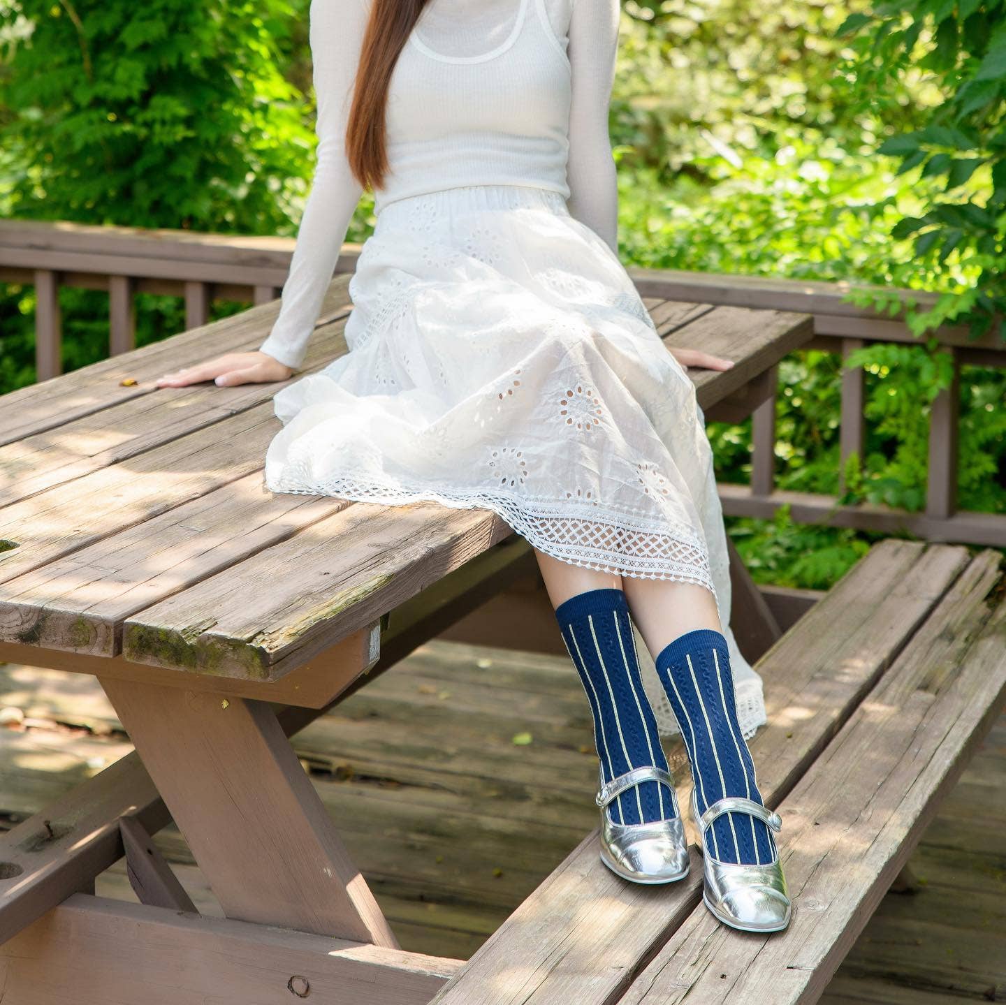 Person wearing white dress and blue socks with silver sandals sitting on a wooden bench outdoors.