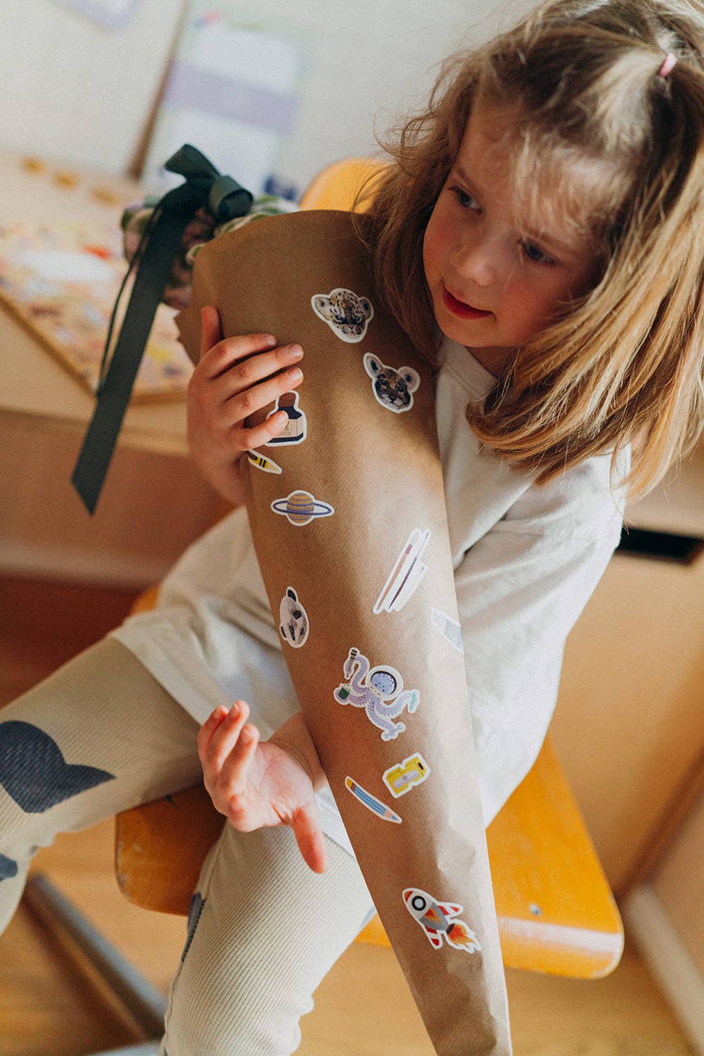 A young girl sitting on a wooden chair, holding up a long brown paper leg sleeve decorated with various cartoon space-themed stickers like planets, rockets, and astronauts. She is wearing a white t-shirt and light-colored pants.