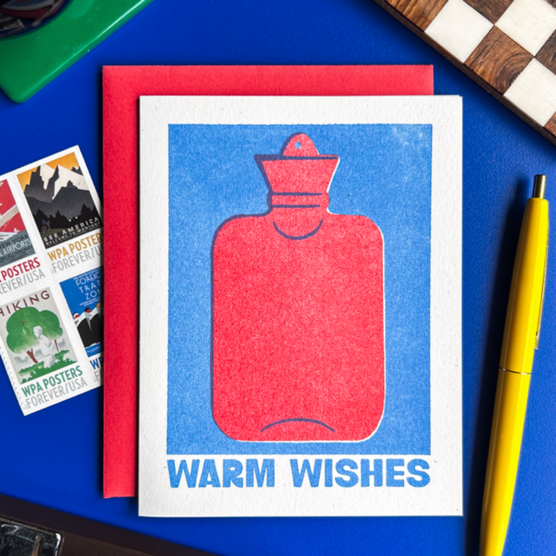 A square greeting card with a red hot water bottle illustration on a blue background and the words 'WARM WISHES' at the bottom, placed on top of a red envelope on a blue tabletop with a yellow pen, postage stamps, and a checkerboard partially visible.