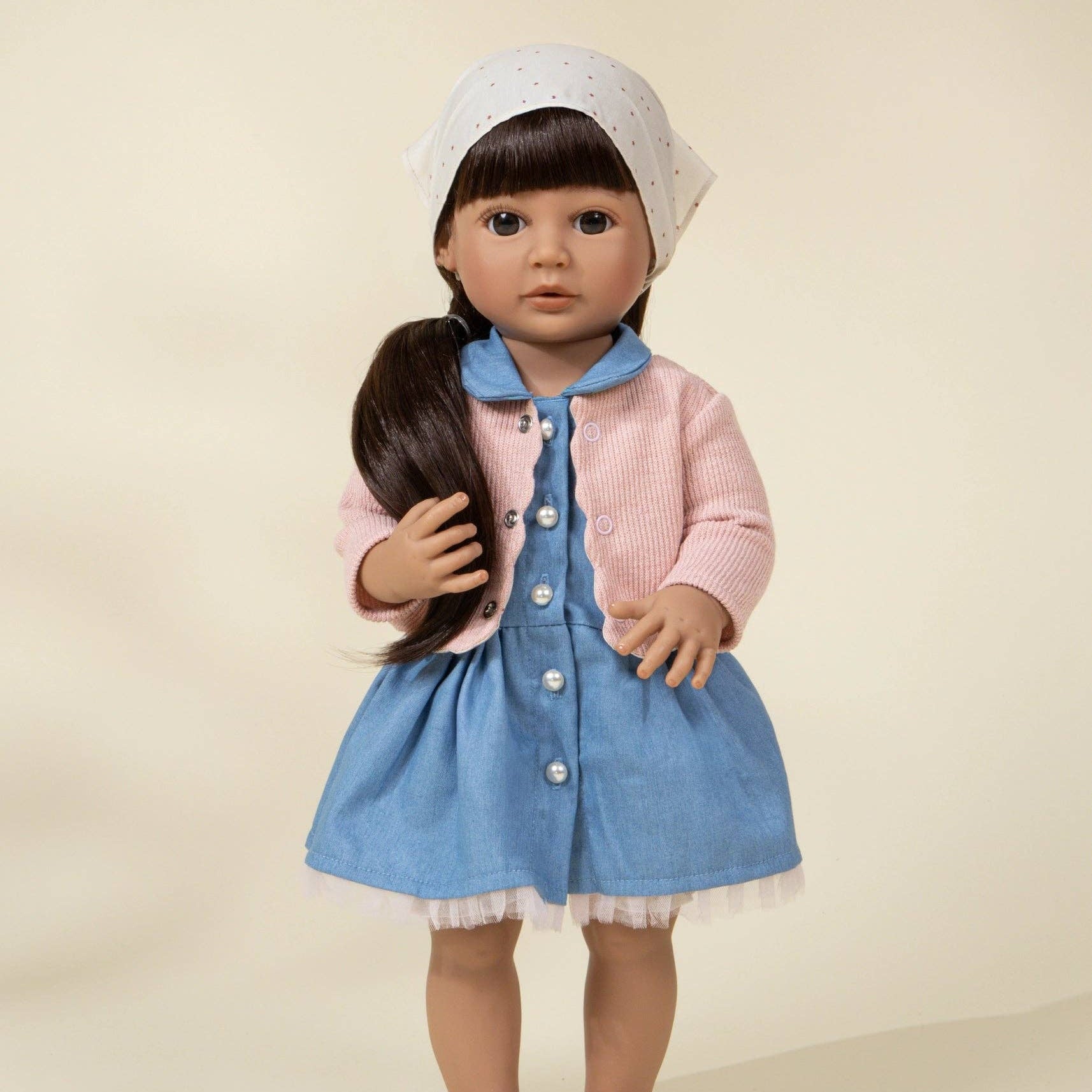 Doll wearing a pink cardigan, blue dress, and white bonnet on a beige background