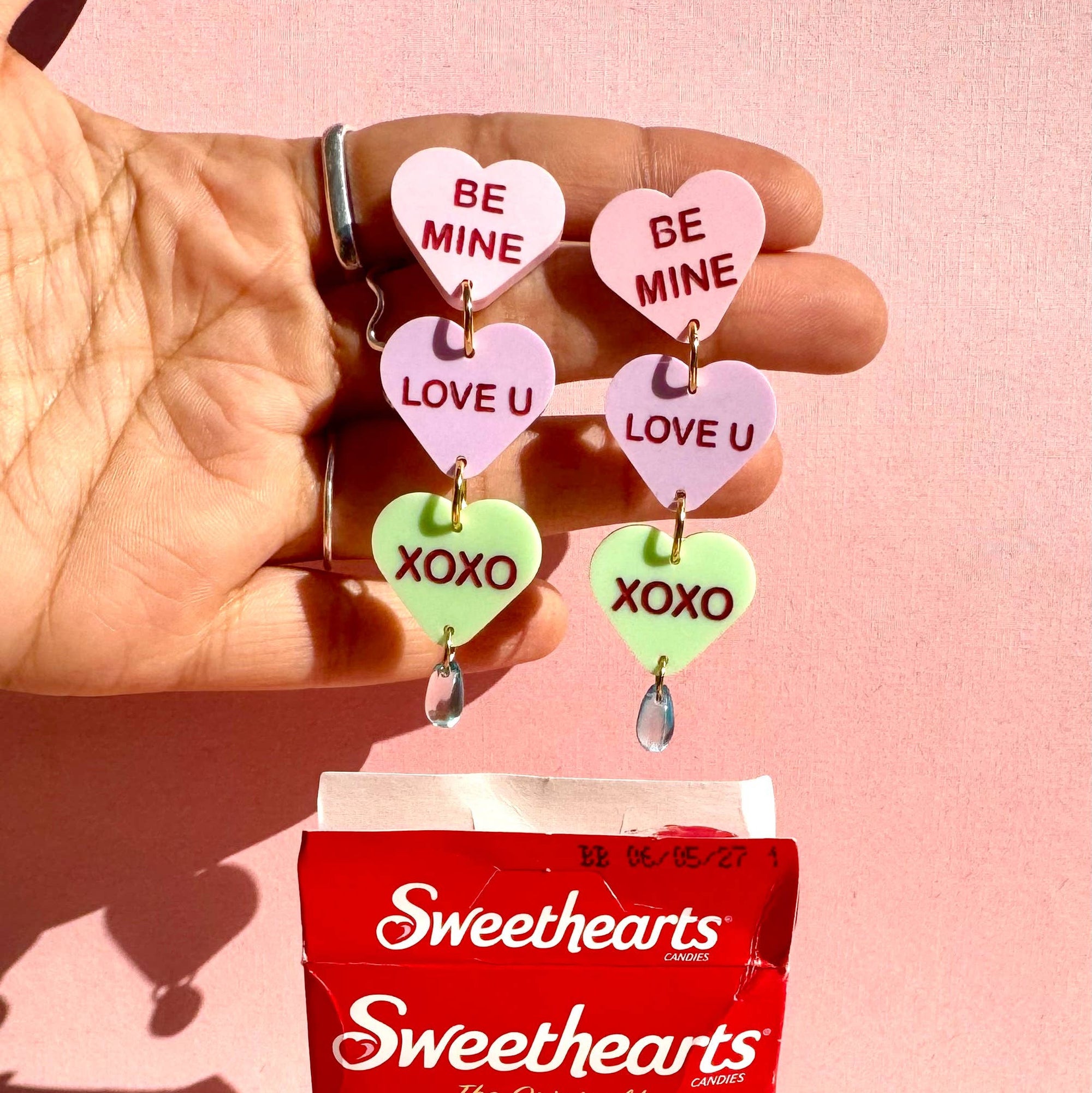 Hand holding Sweethearts candy with heart-shaped charms on a pink background