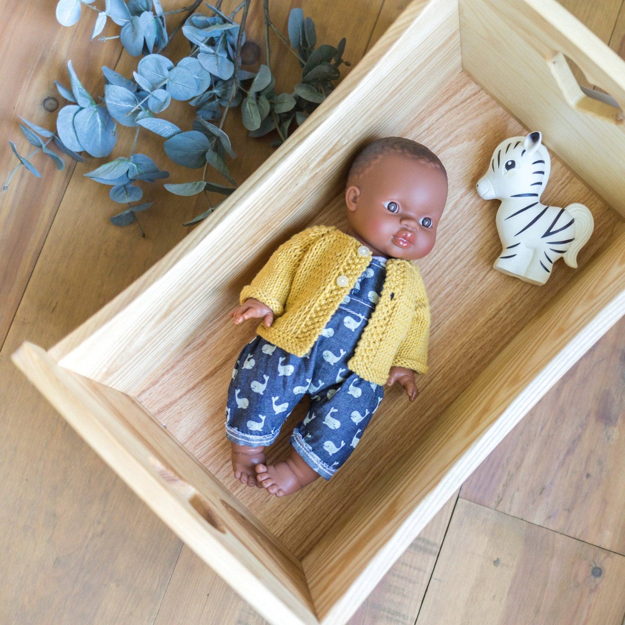 Baby doll in a yellow sweater and blue pants with a zebra toy in a wooden box on a wooden floor.