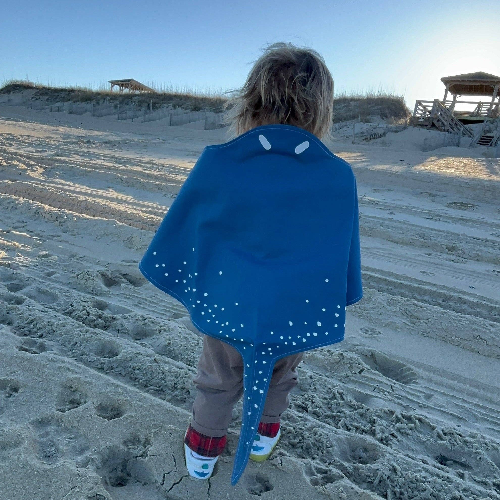 Person walking on a sandy beach with a blue towel draped over their shoulders.