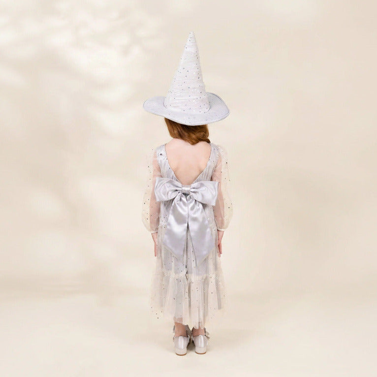 A child viewed from behind wearing a white witch-style pointed hat with small silver embellishments and a white tulle dress with a large satin bow at the back, standing against a warm cream studio background.