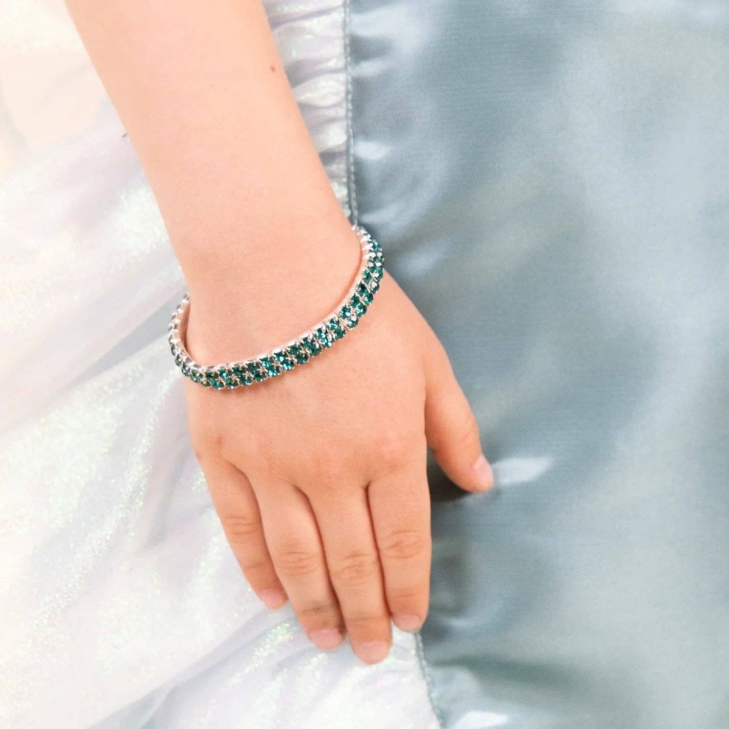 A close-up of a person's hand and wrist wearing a thin silver-toned tennis bracelet with green gemstones, set against a light blue satin fabric background.