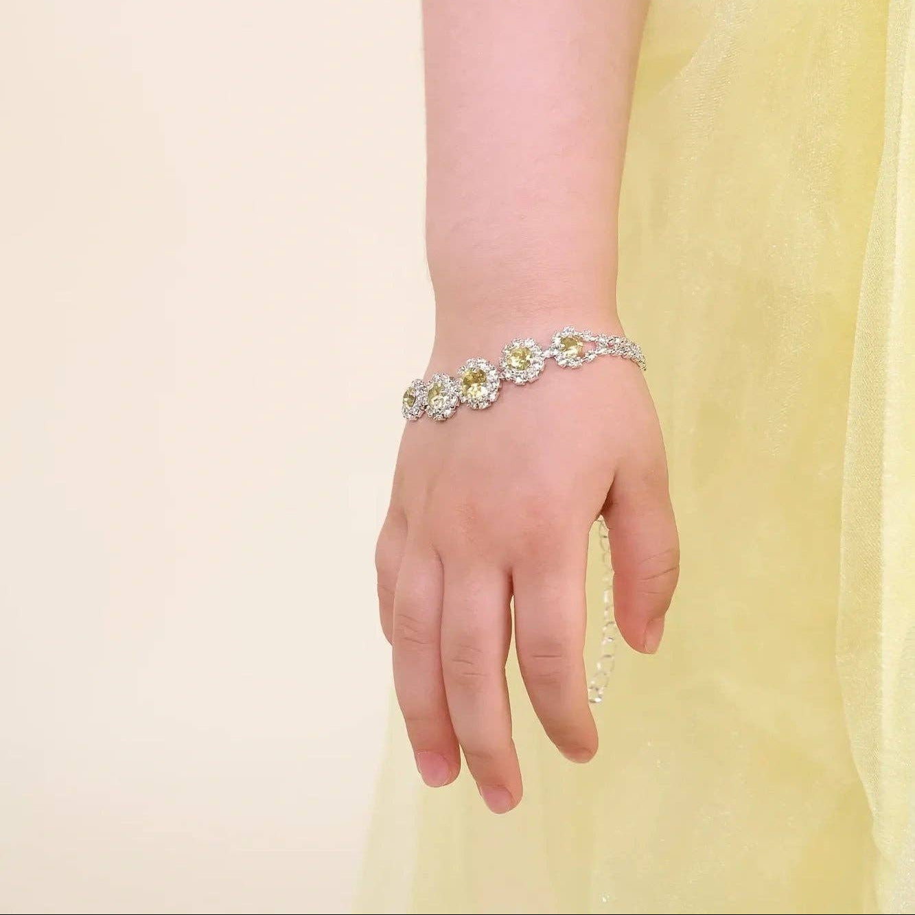 A close-up of a child's hand and wrist wearing a delicate silver-toned bracelet adorned with small white and yellow floral details, set against a soft cream background and a pale yellow dress.