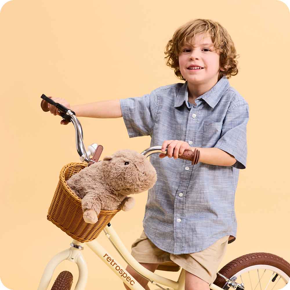 Child on a bicycle with a basket containing a plush toy against a yellow background