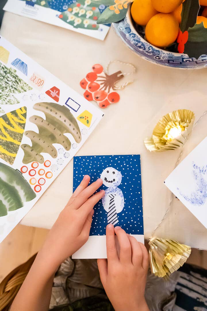 A child's hands are shown assembling a handmade snowman craft on a blue card, surrounded by colorful craft supplies, paper fans, and warm string lights on a light wooden table.