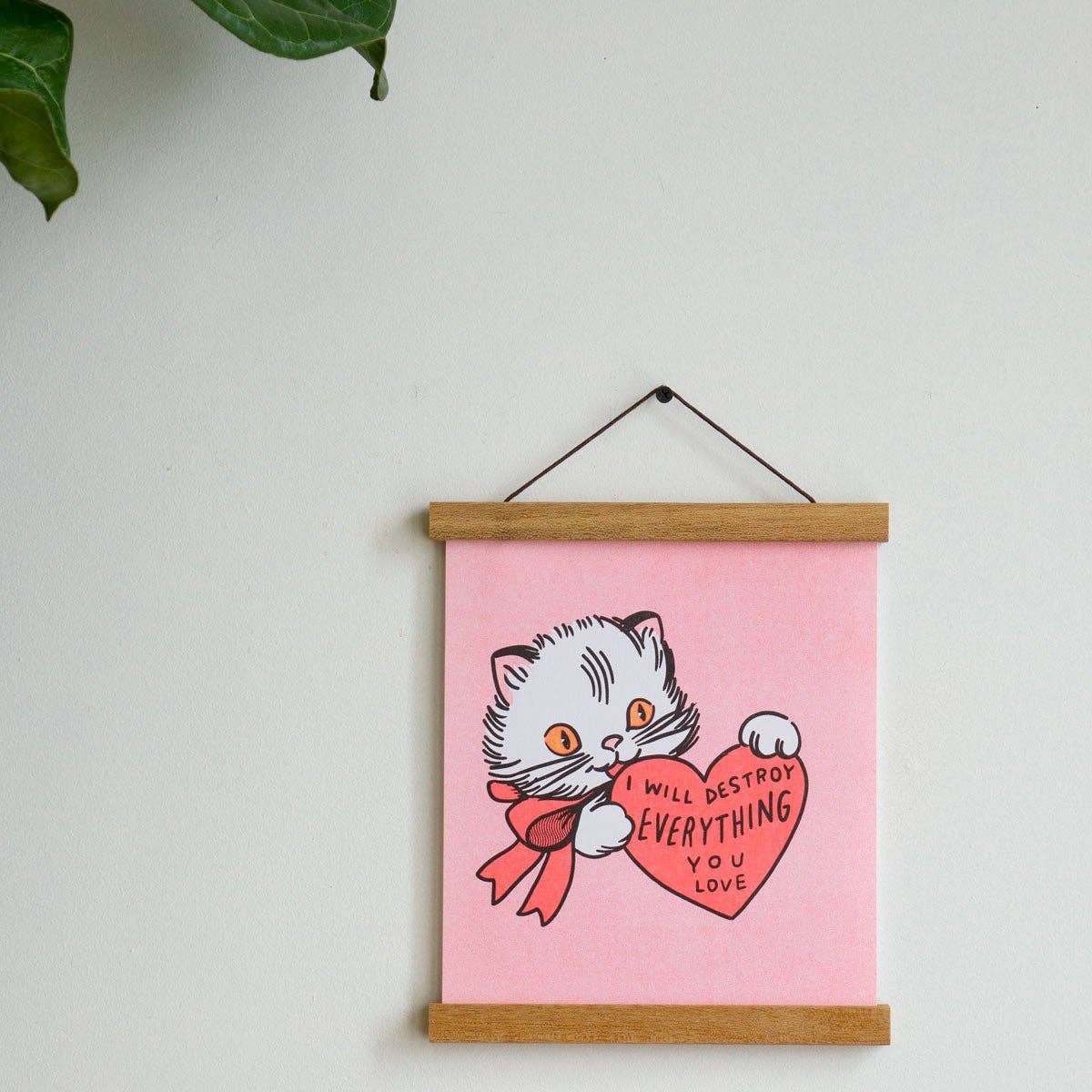 Illustration of a cat holding a heart with text on a pink background, hanging on a white wall.