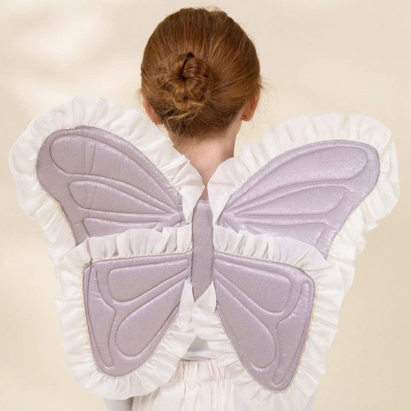 A child viewed from behind wearing a butterfly wing costume accessory with large lavender quilted wings and white ruffled trim, paired with cream clothing against a soft beige studio background.
