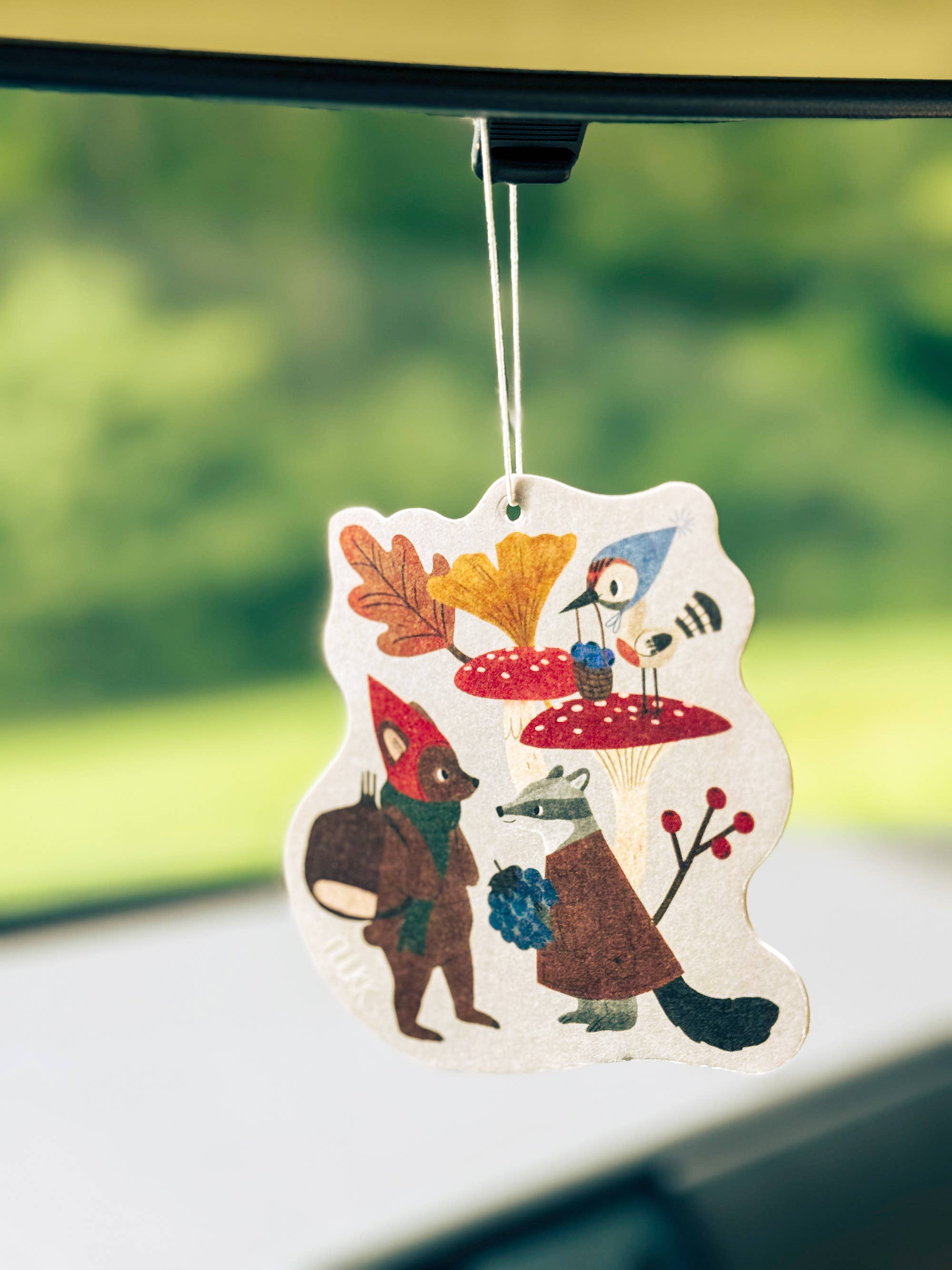 A whimsical forest-themed car air freshener hanging from a black car window clip. The die-cut white paper ornament features painted woodland characters including a fox, a bird, a mushroom, and colorful autumn leaves, set against a blurred green outdoor background.