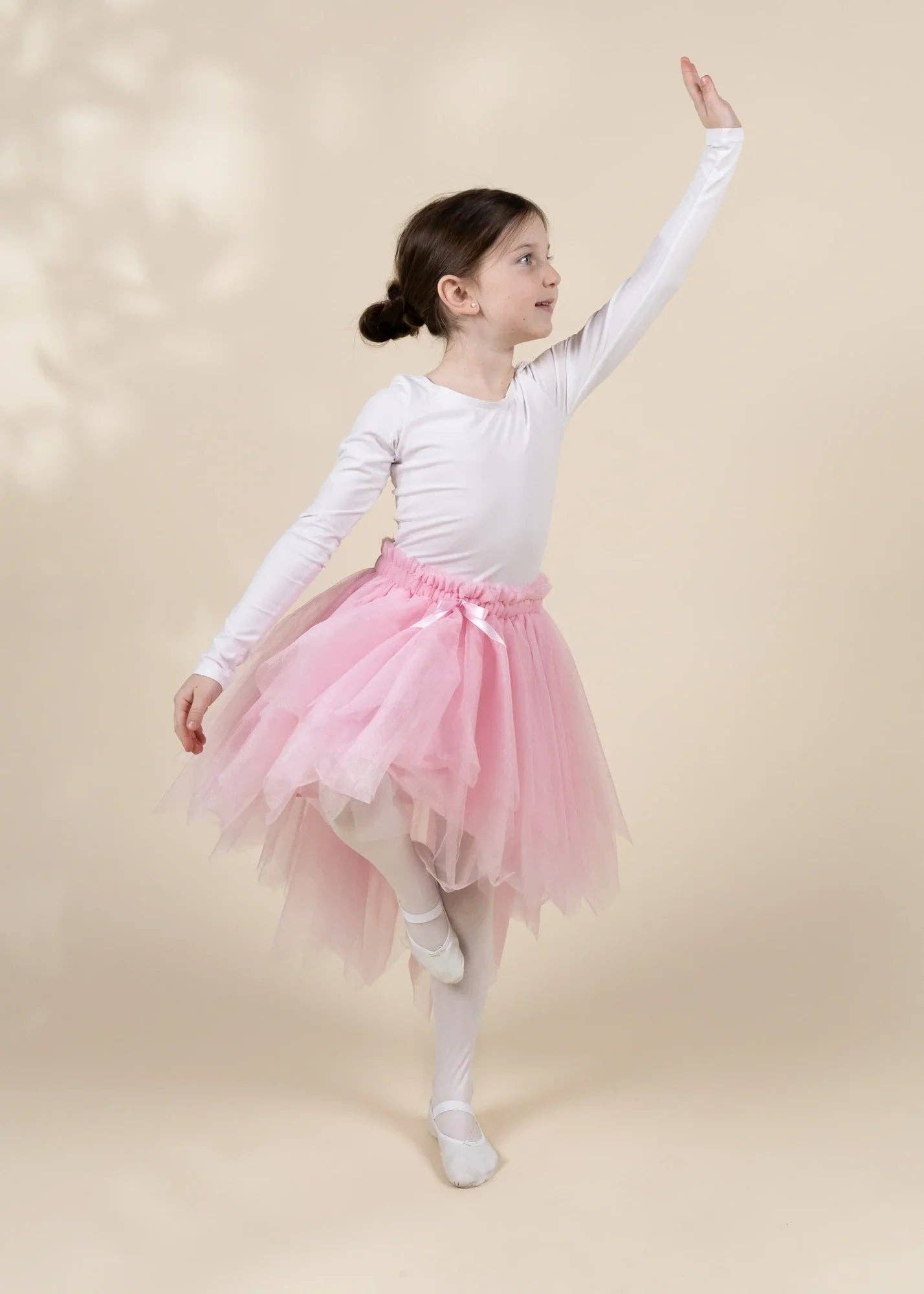 A young girl dancer posing in a white long-sleeve leotard and a pink tulle tutu skirt against a warm beige background.