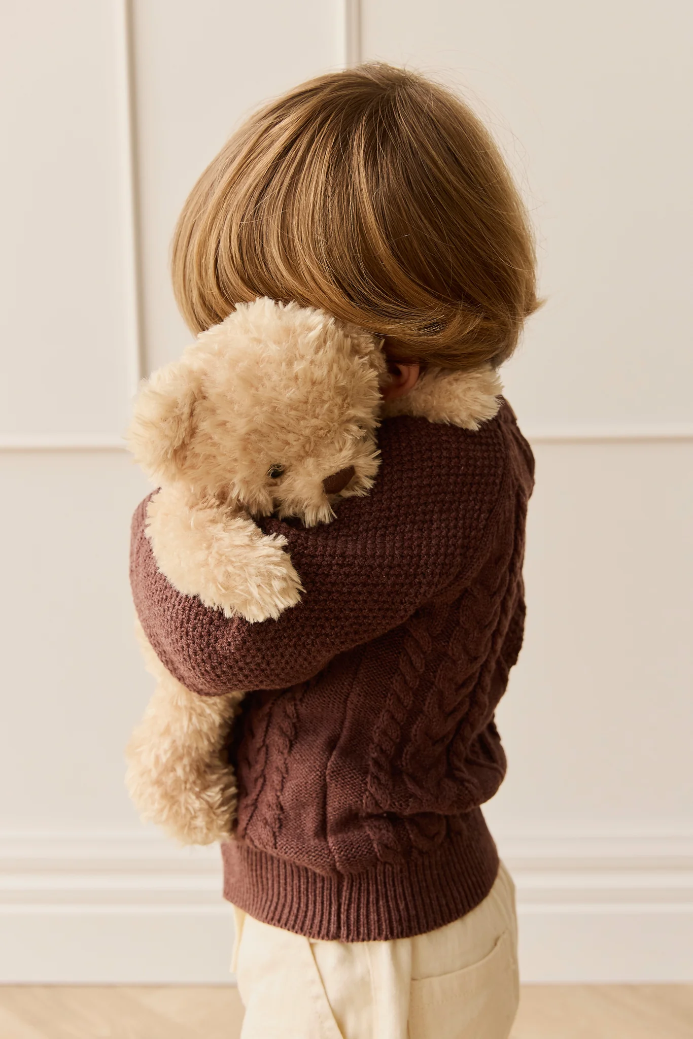 Child wearing a brown sweater holding a teddy bear against a white wall.