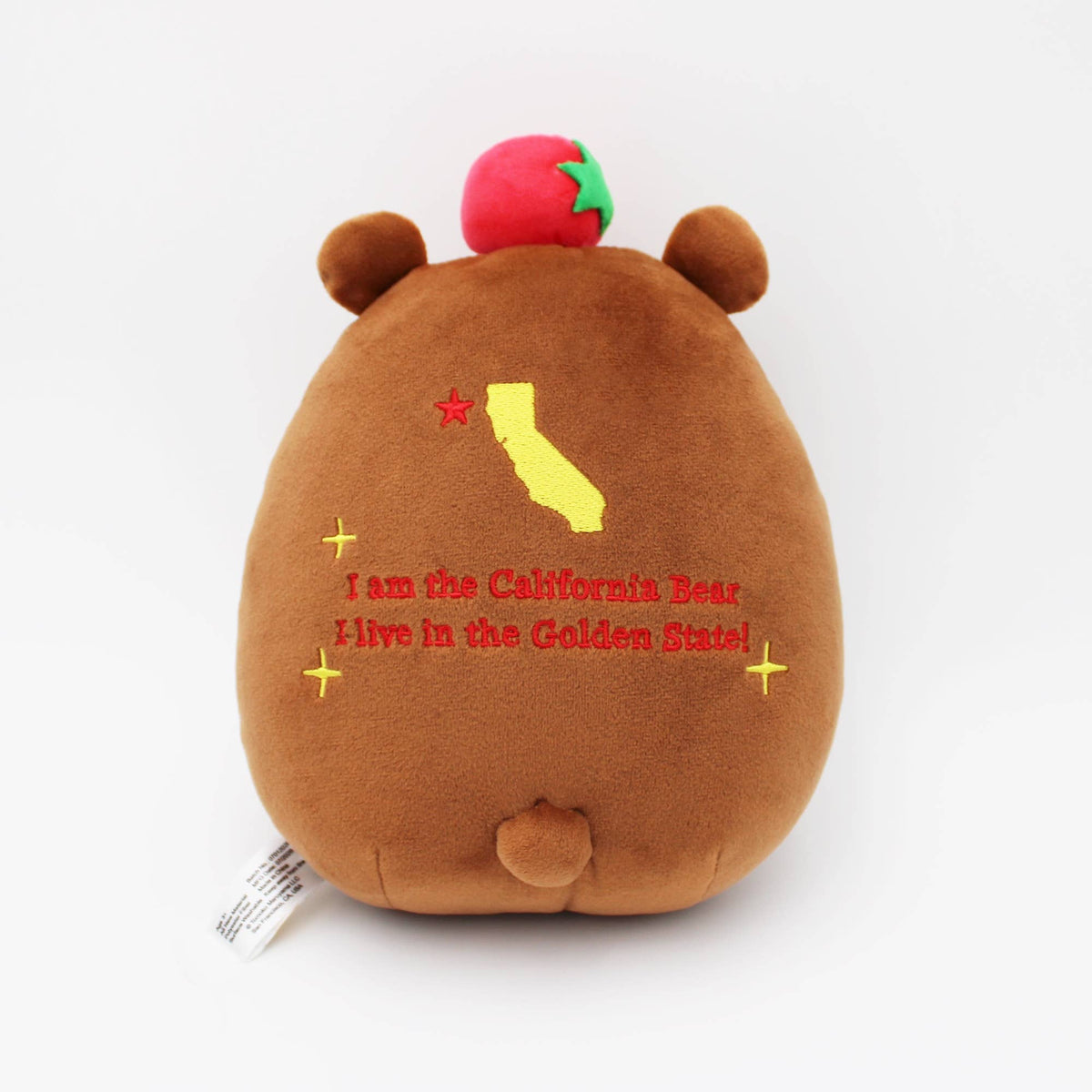 A brown plush bear toy shown from the back, featuring a red strawberry on its head, a yellow California state silhouette, and red and yellow embroidered text reading 'I am the California Bear. I live in the Golden State!'.