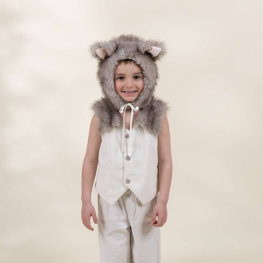 A young child modeling a wolf costume consisting of a gray faux-fur hood with ears, a cream vest, matching cream pants, and gray socks, set against a warm beige studio background.
