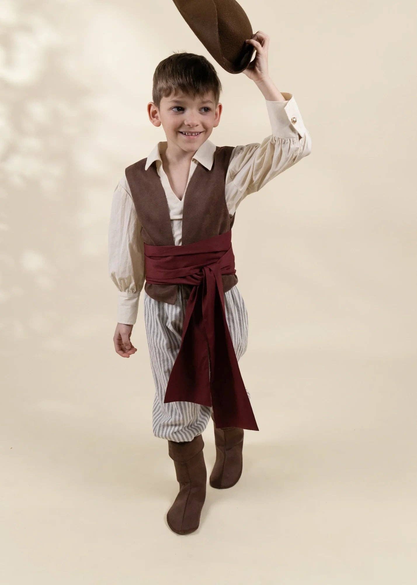 A young boy modeling a pirate costume against a warm beige background. The costume includes a brown vest, a cream-colored shirt, a dark red sash, striped pants, and brown boots, with the boy holding a brown hat above his head.