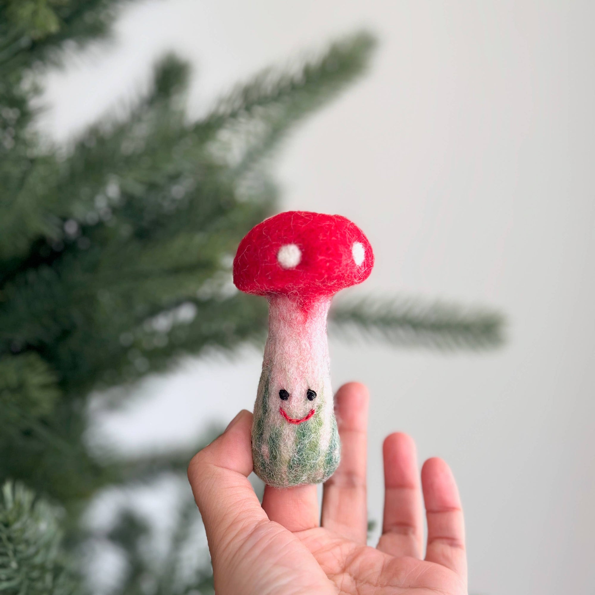 Hand holding a small felted mushroom toy with a red cap and white spots against a blurred green background.