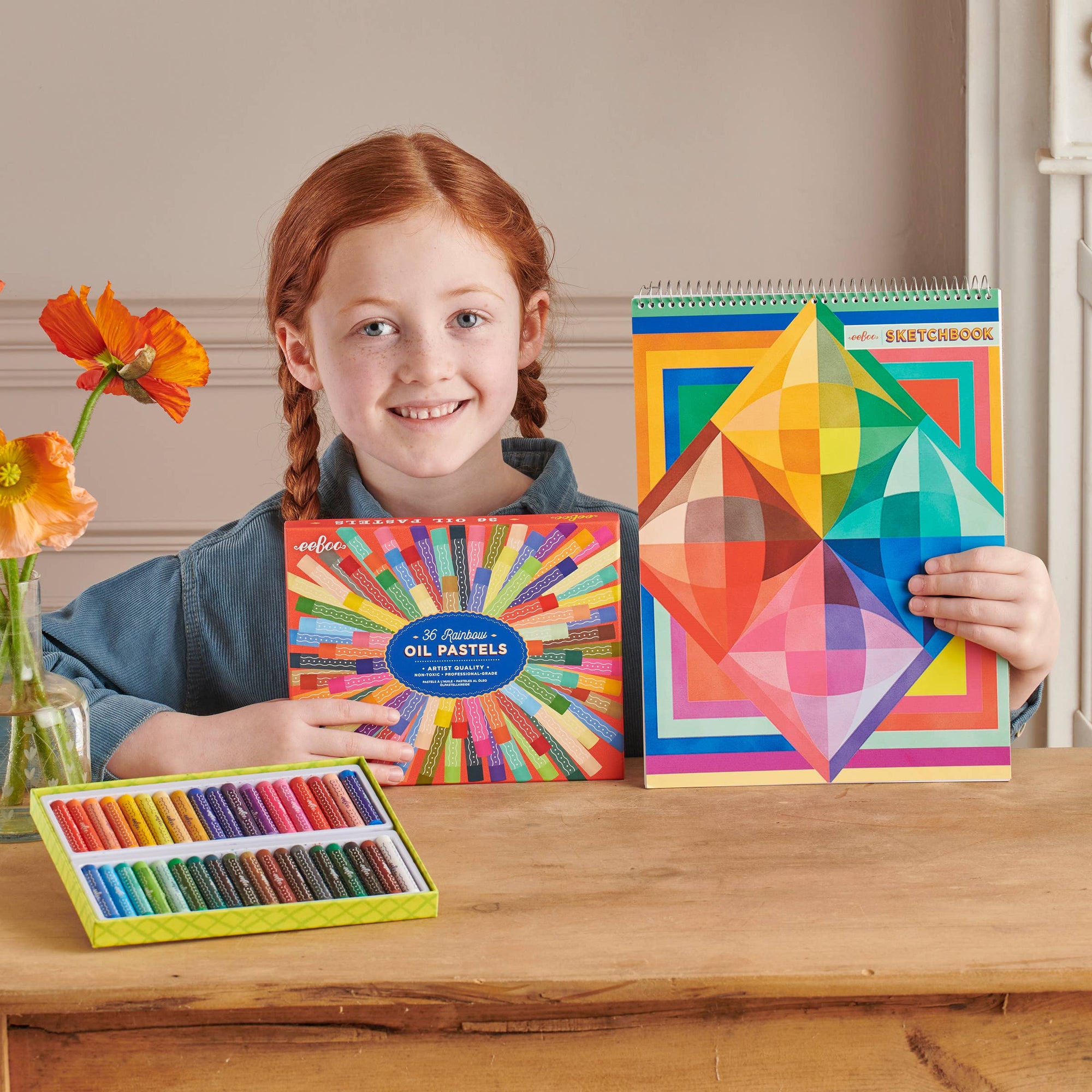 Girl holding art supplies including oil pastels and a colorful notebook on a wooden table.