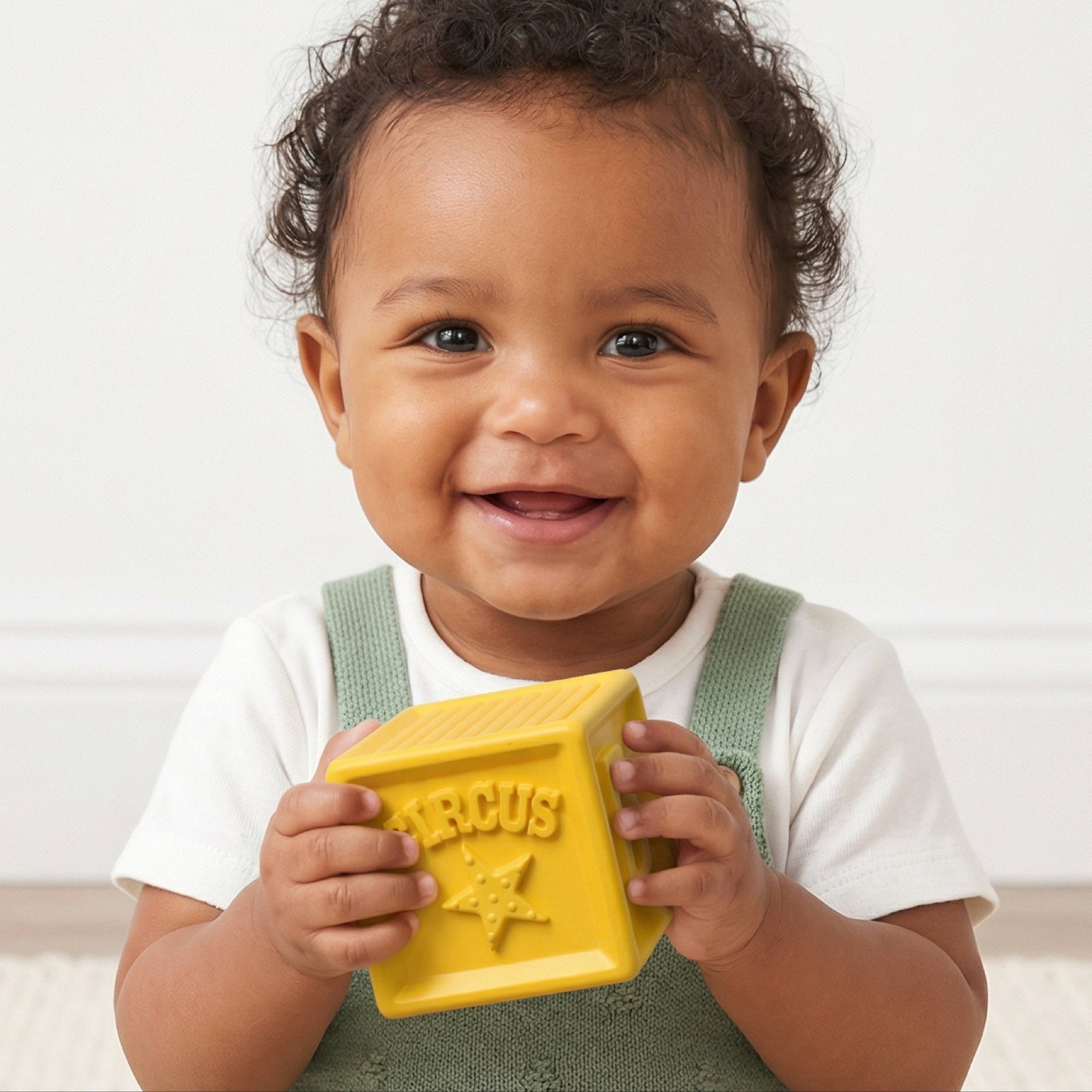 A smiling toddler in a white t-shirt and green overalls holds a bright yellow block toy embossed with the word 'CIRCUS' and a star. The child is photographed against a clean, off-white background.