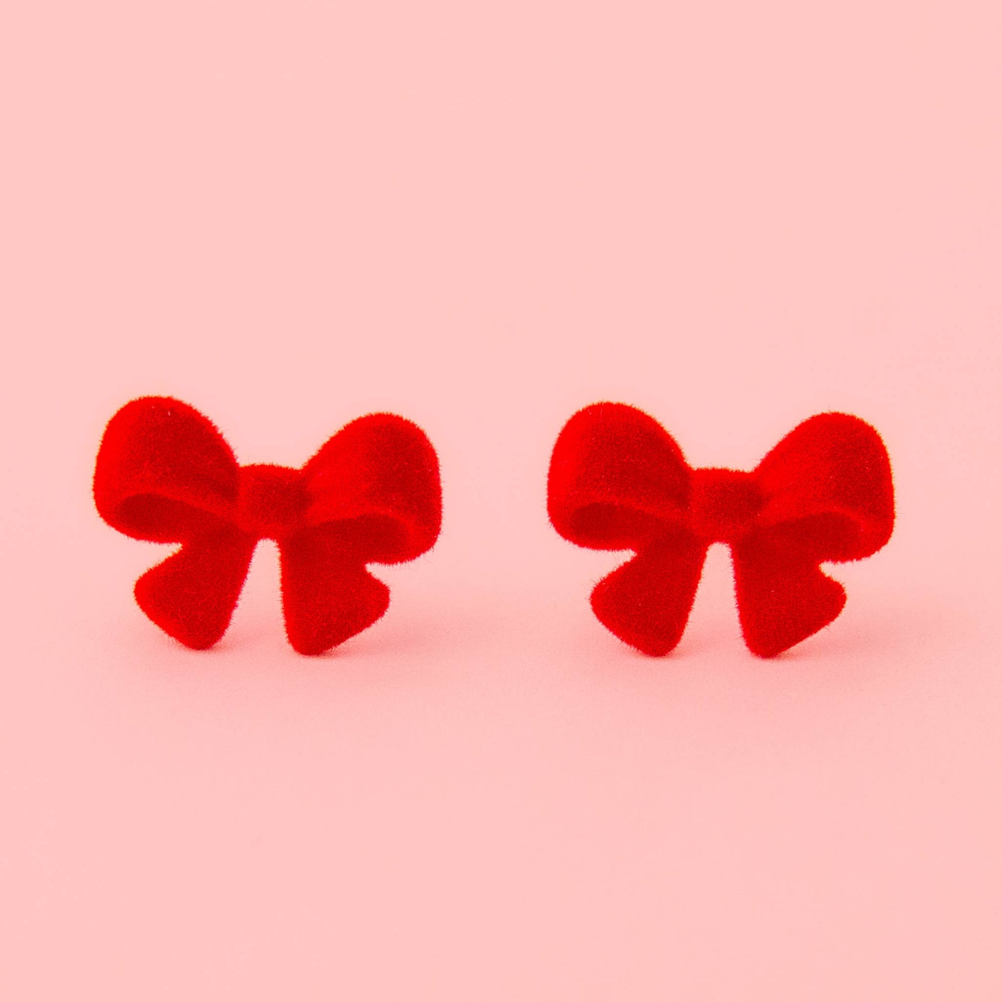 A pair of red velvet bow stud earrings on a solid light pink background.