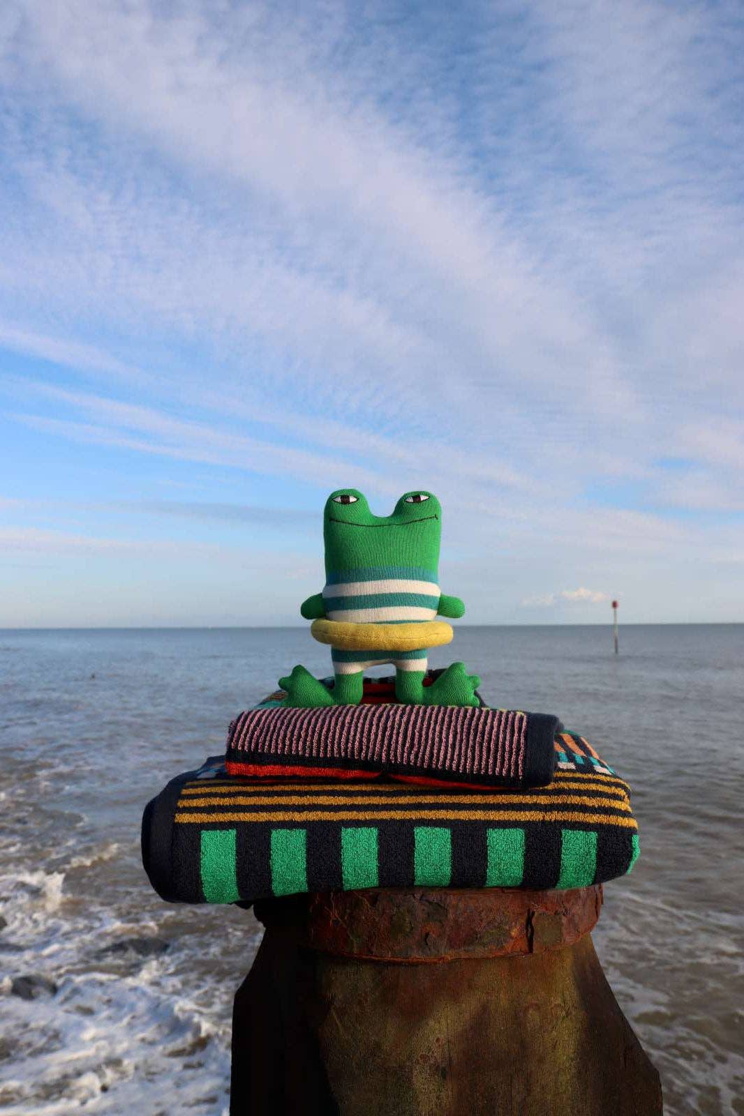 A green plush frog wearing a striped sweater sits on top of folded striped knit blankets, placed on a weathered wooden post by the ocean with a blue sky and clouds in the background.