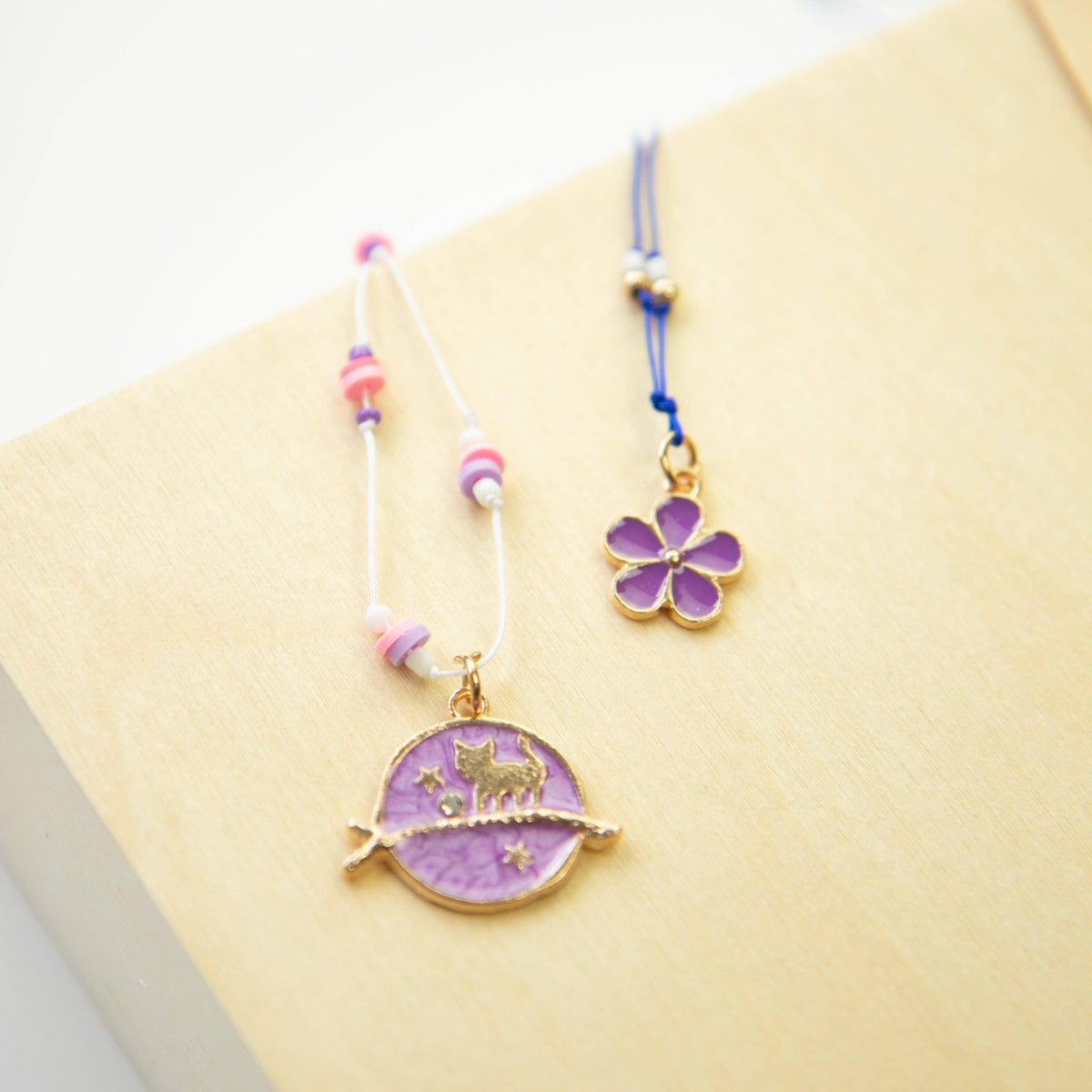 Two delicate jewelry charms displayed on a light wood surface. One charm features a purple crescent moon and cat pendant with gold details, accented by pink and white beads on a white cord.