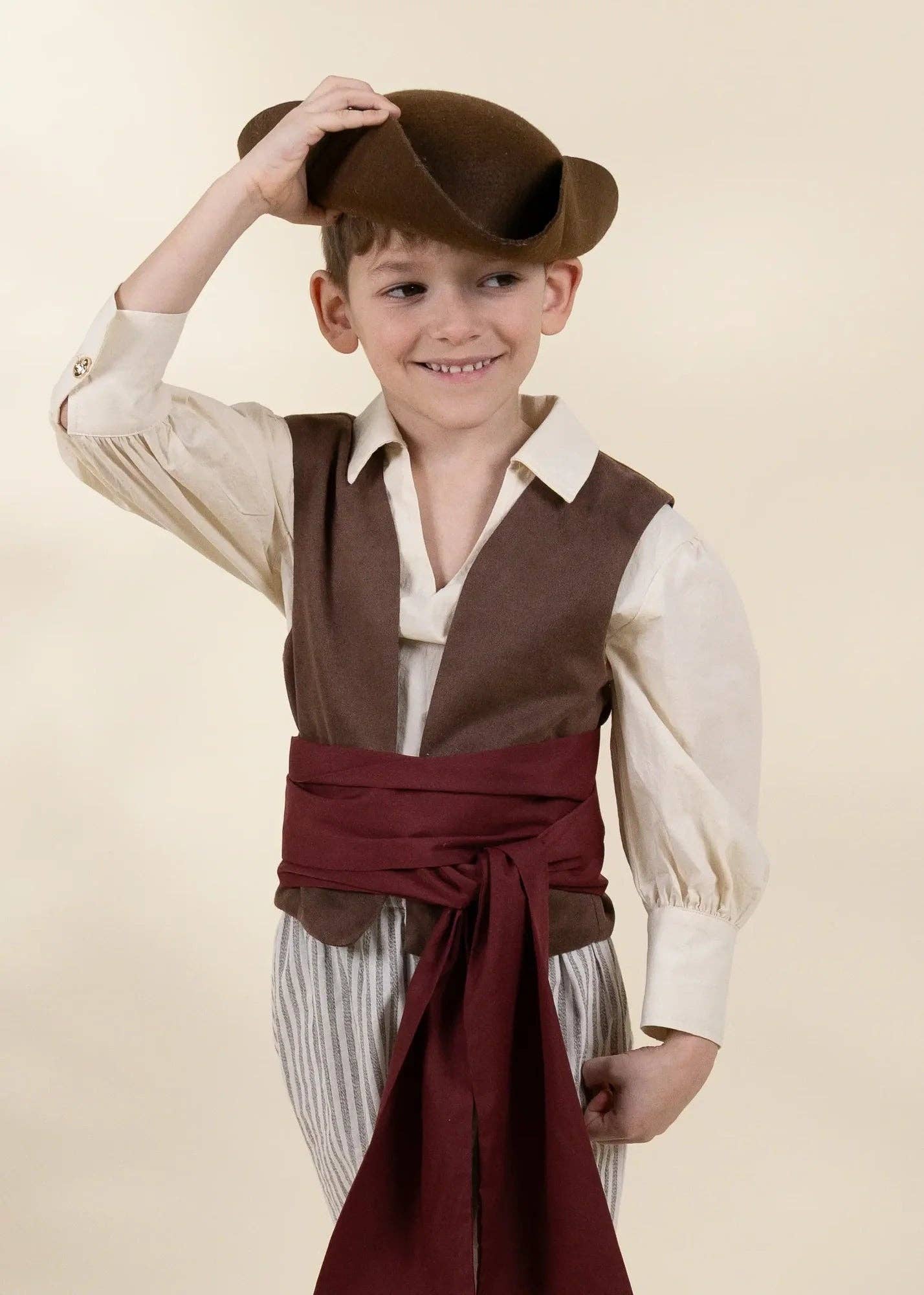 A young boy modeling a pirate costume consisting of a brown hat, brown vest, cream shirt, and a dark red sash, set against a solid beige background.