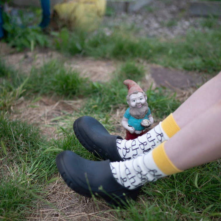 Person wearing black shoes and patterned socks with a garden gnome in the grass.