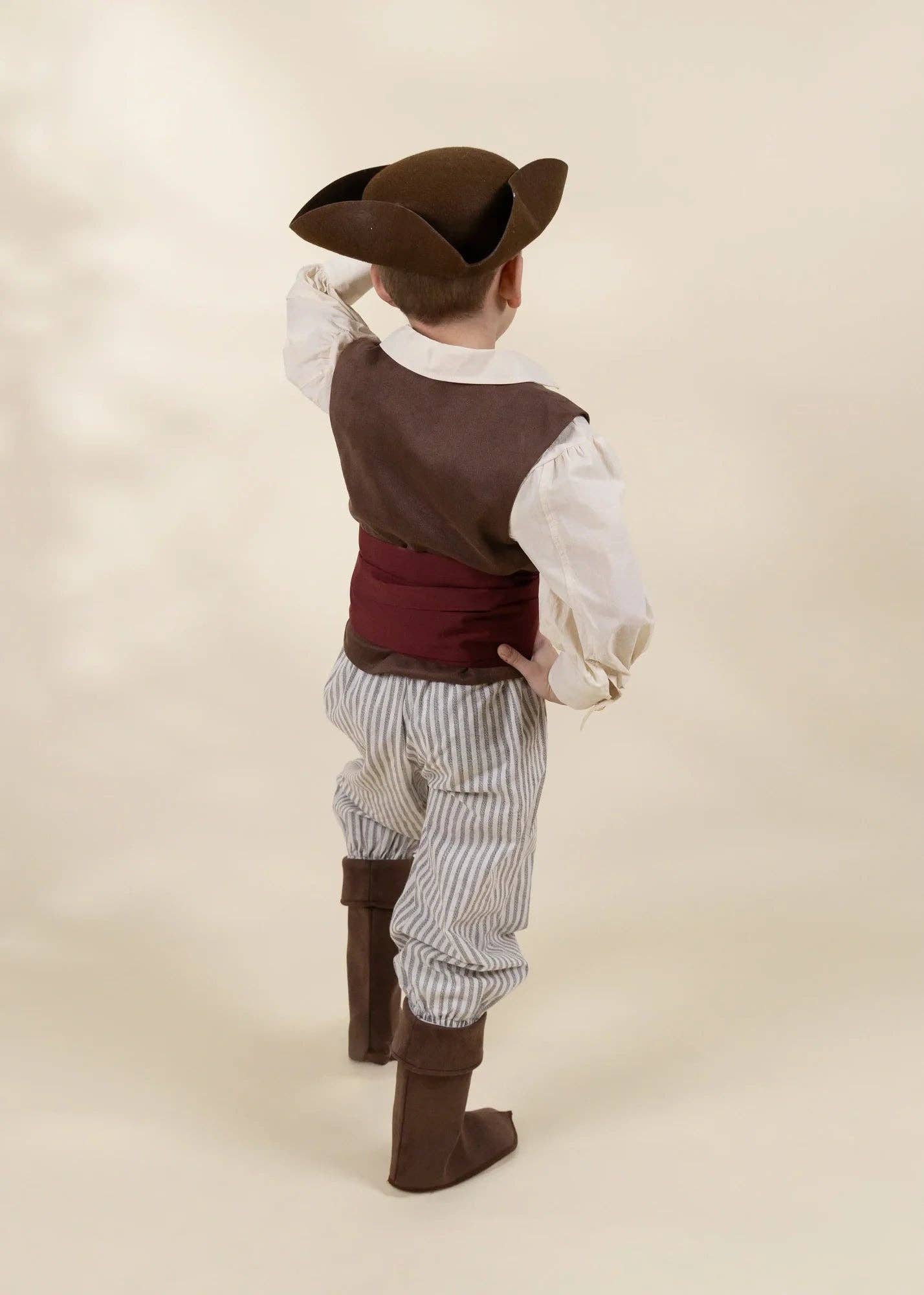 A child wearing a pirate costume, shown from the back. The costume includes a brown tricorn hat, a brown vest over a white shirt, a dark red waist sash, vertically striped gray and white pants, and brown boots, set against a plain beige studio background.