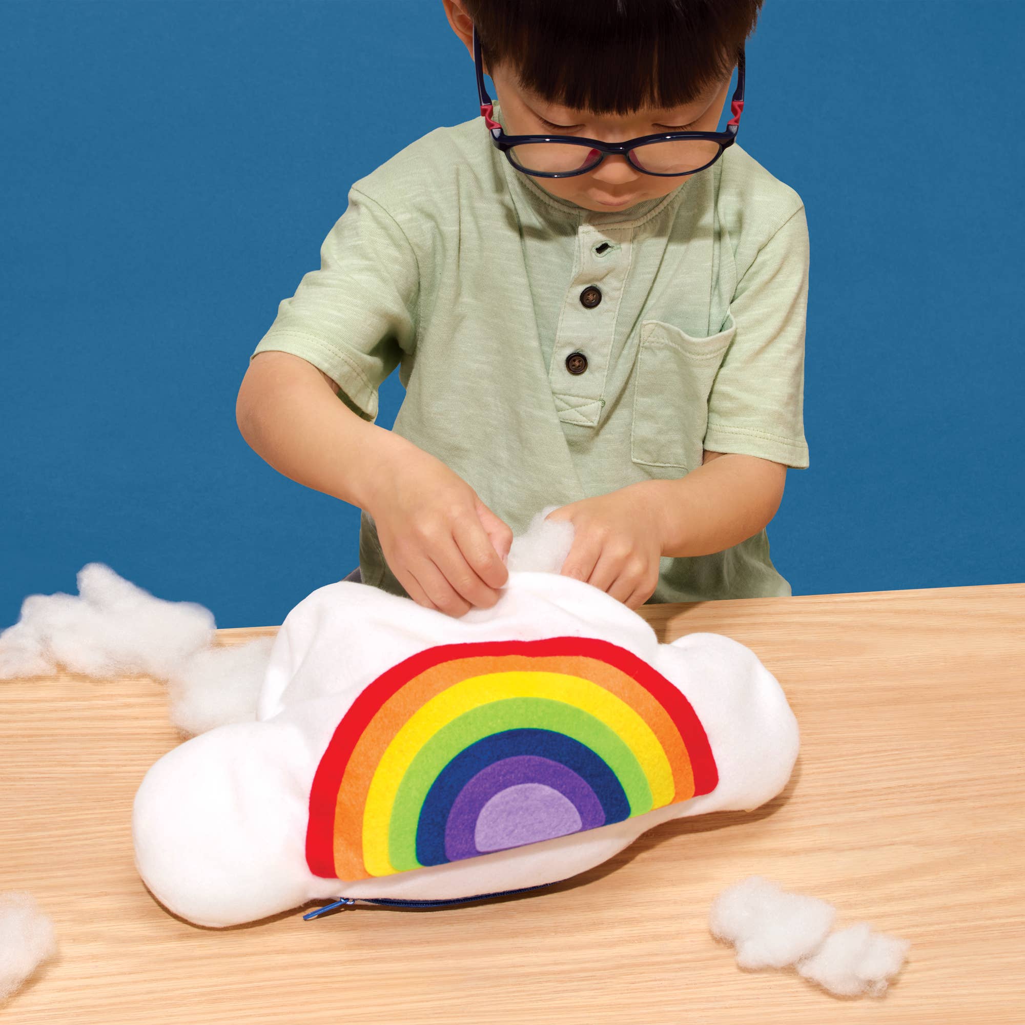 A child wearing glasses and a light green shirt is stuffing a white cloud-shaped zippered pouch with white faux fur. The pouch features a colorful rainbow design on the front, set on a light wood table against a solid blue background.