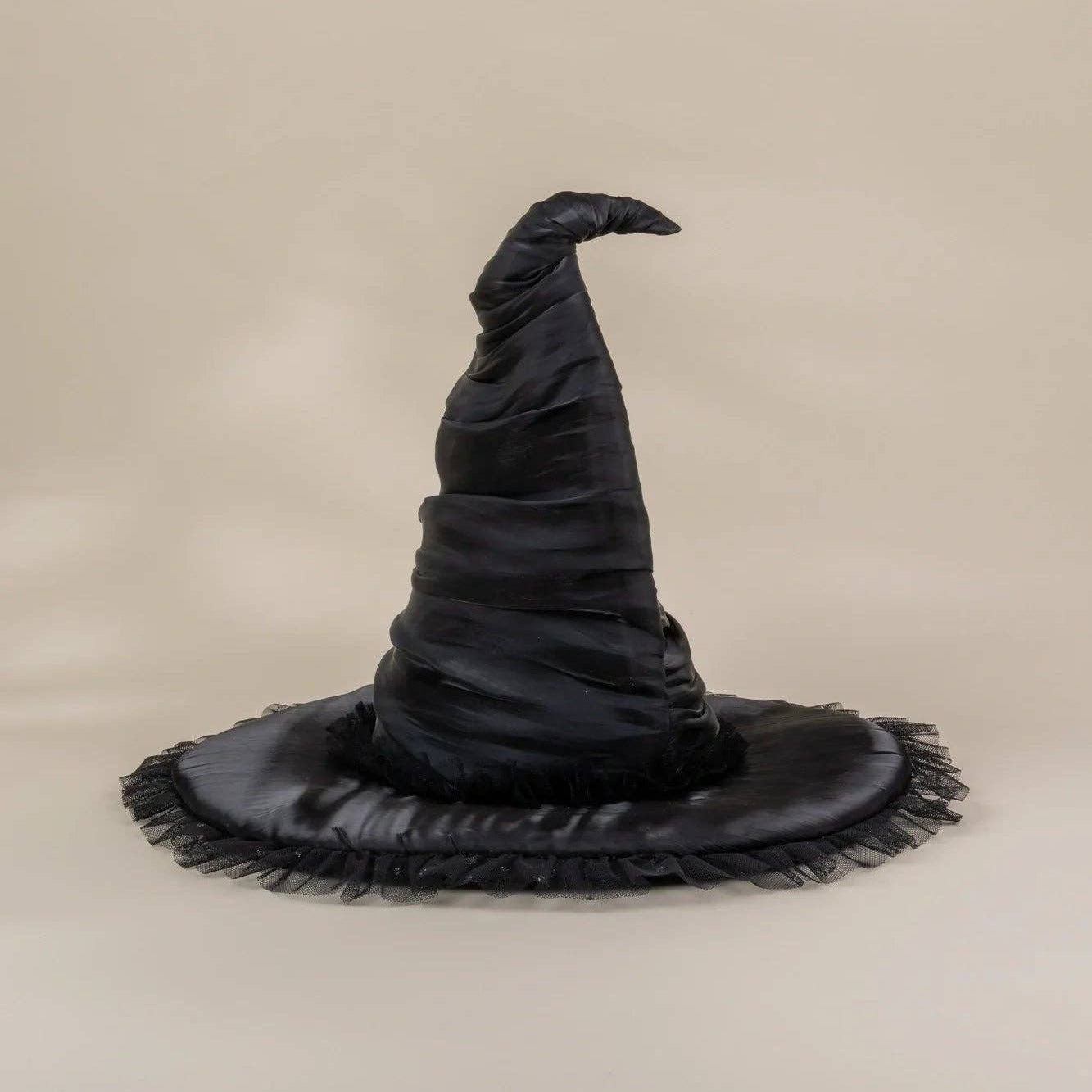 A black satin witch hat with a wide, frayed brim, photographed against a solid beige studio background.