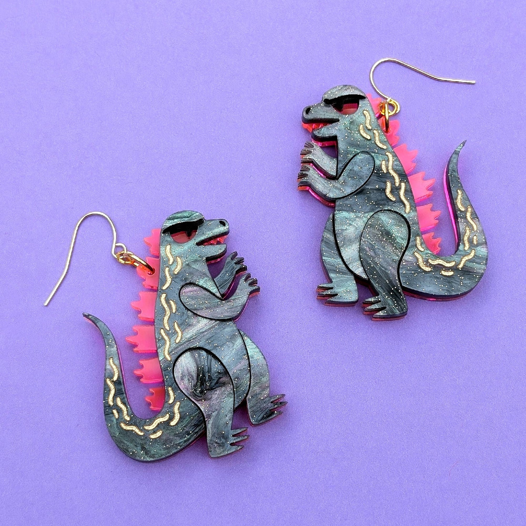 Dinosaur-shaped earrings on a purple background with 'Sylvia Draws!' branding.