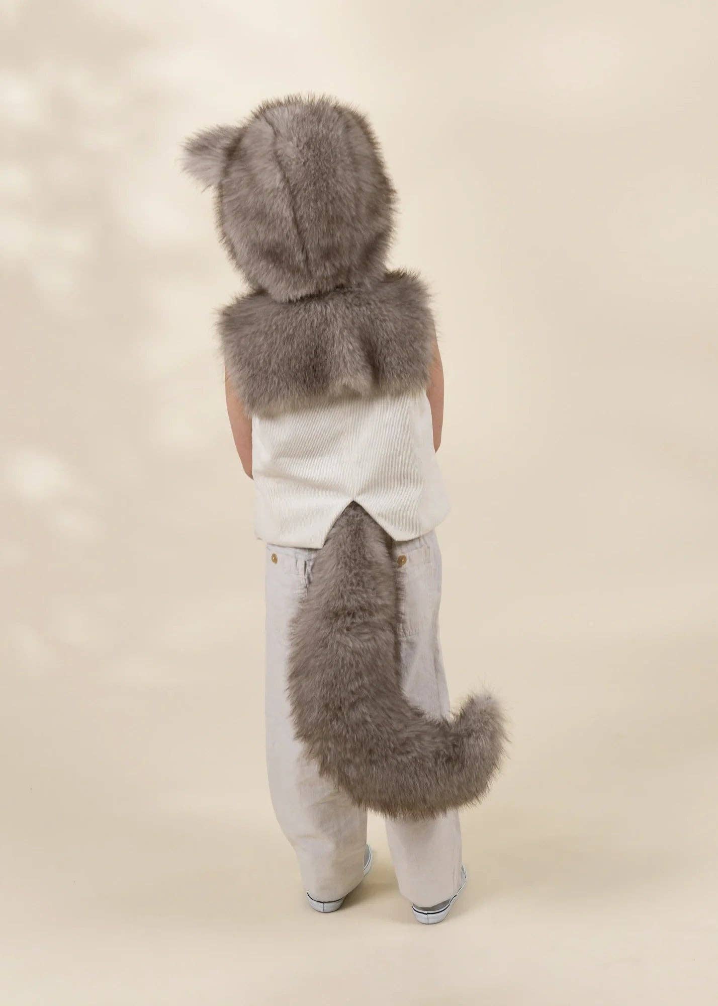 A child shown from the back wearing a gray faux-fur hooded costume with a matching tail, over a white top and white pants, against a plain beige studio background.