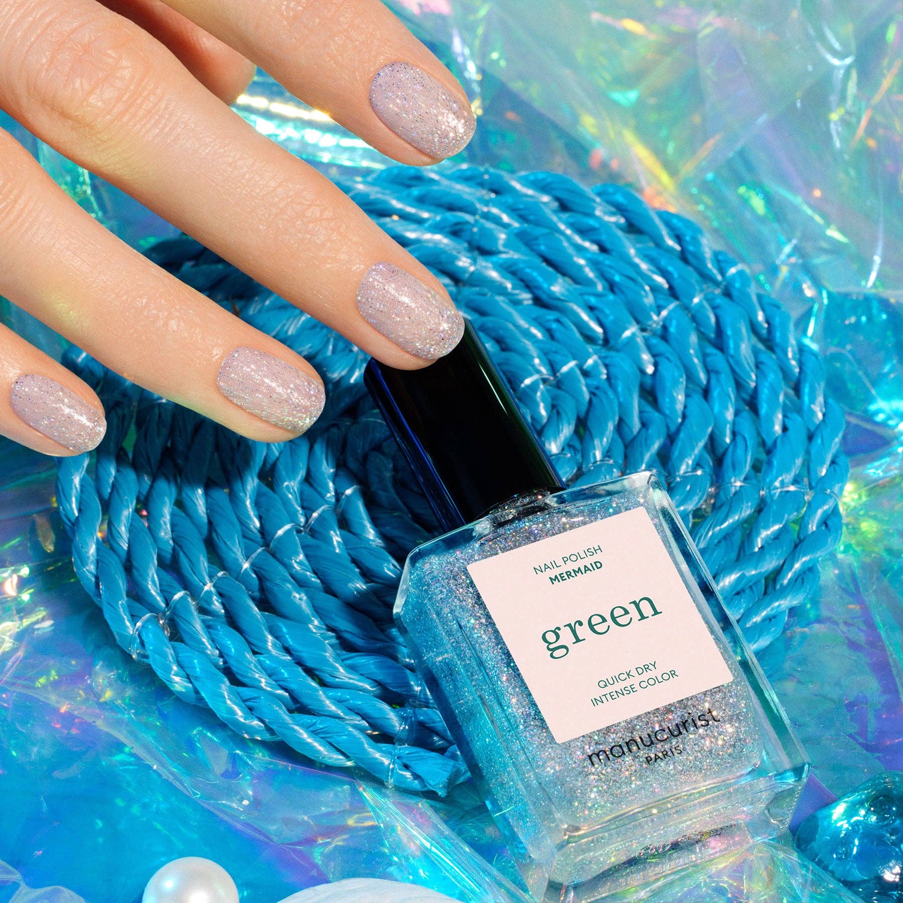 Manicured hand with glittery nails holding a bottle of 'green' nail polish against a blue textured background.