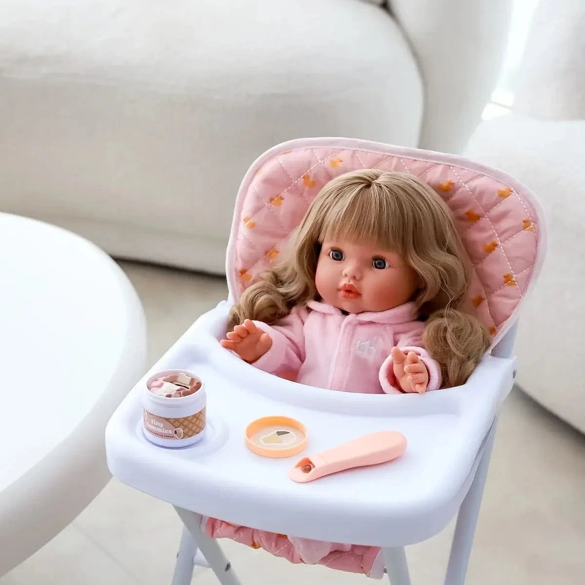 Doll in a high chair with pink cushion and food toys on a light-colored floor.