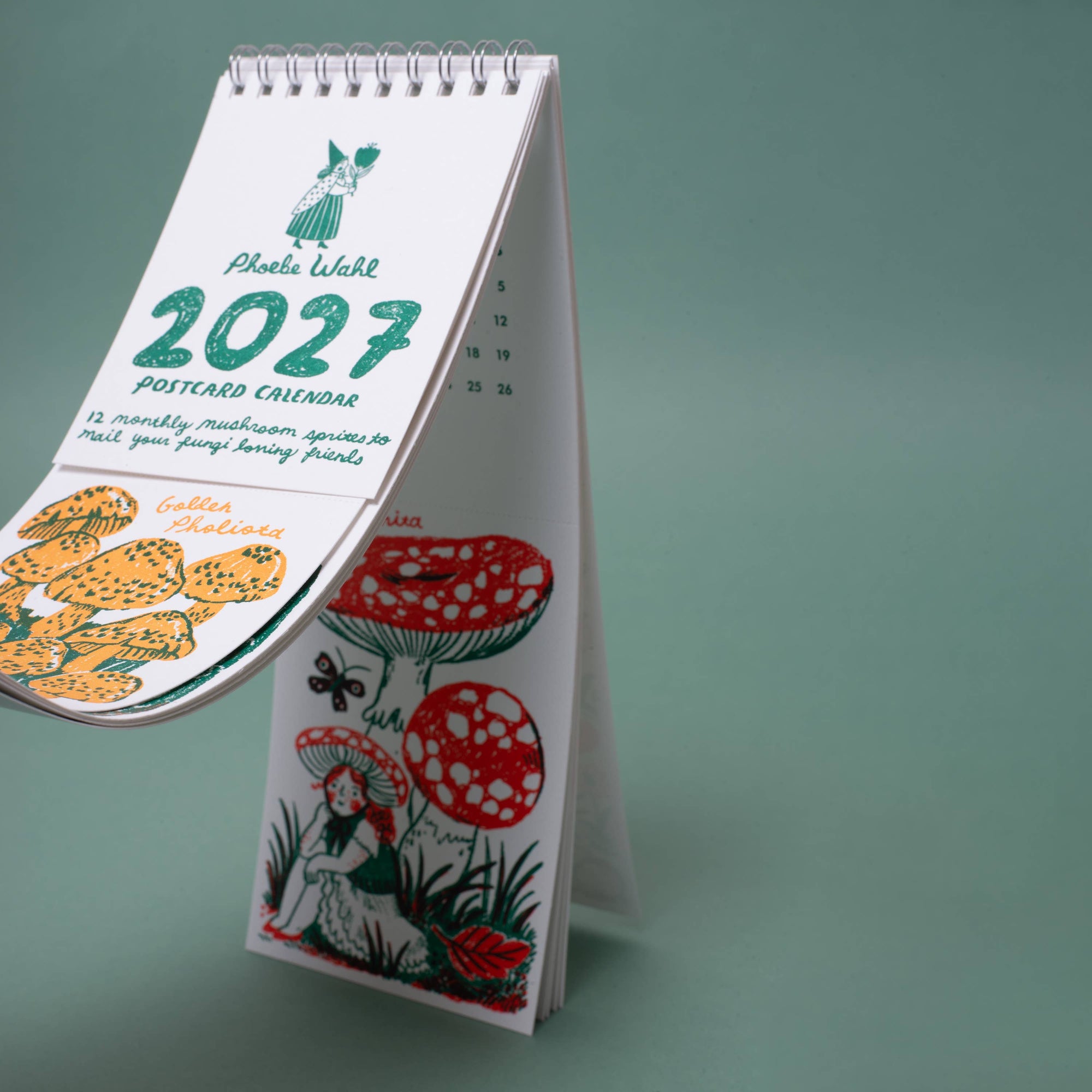 A spiral-bound 2027 postcard calendar  stands open on a stand against a muted green background. The calendar pages feature whimsical illustrations, including mushrooms, flowers, a butterfly, and a fairy.