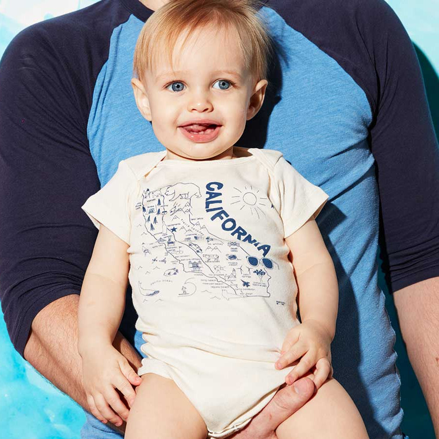 Baby wearing a 'California' onesie sitting on a person's lap with a blue background