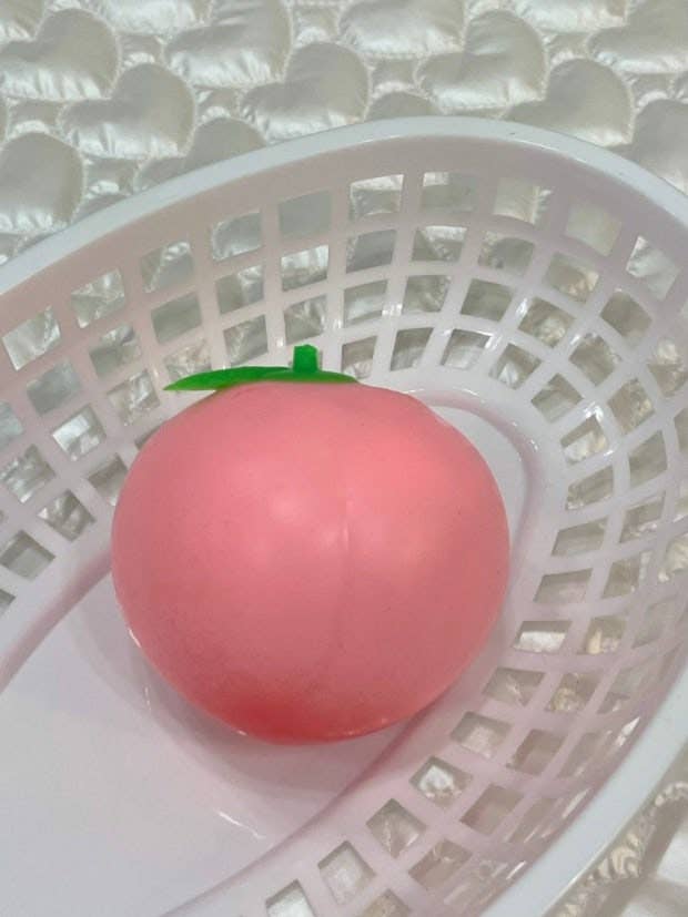 A pink peach-shaped squishy toy with a small green stem, resting inside a white plastic basket.