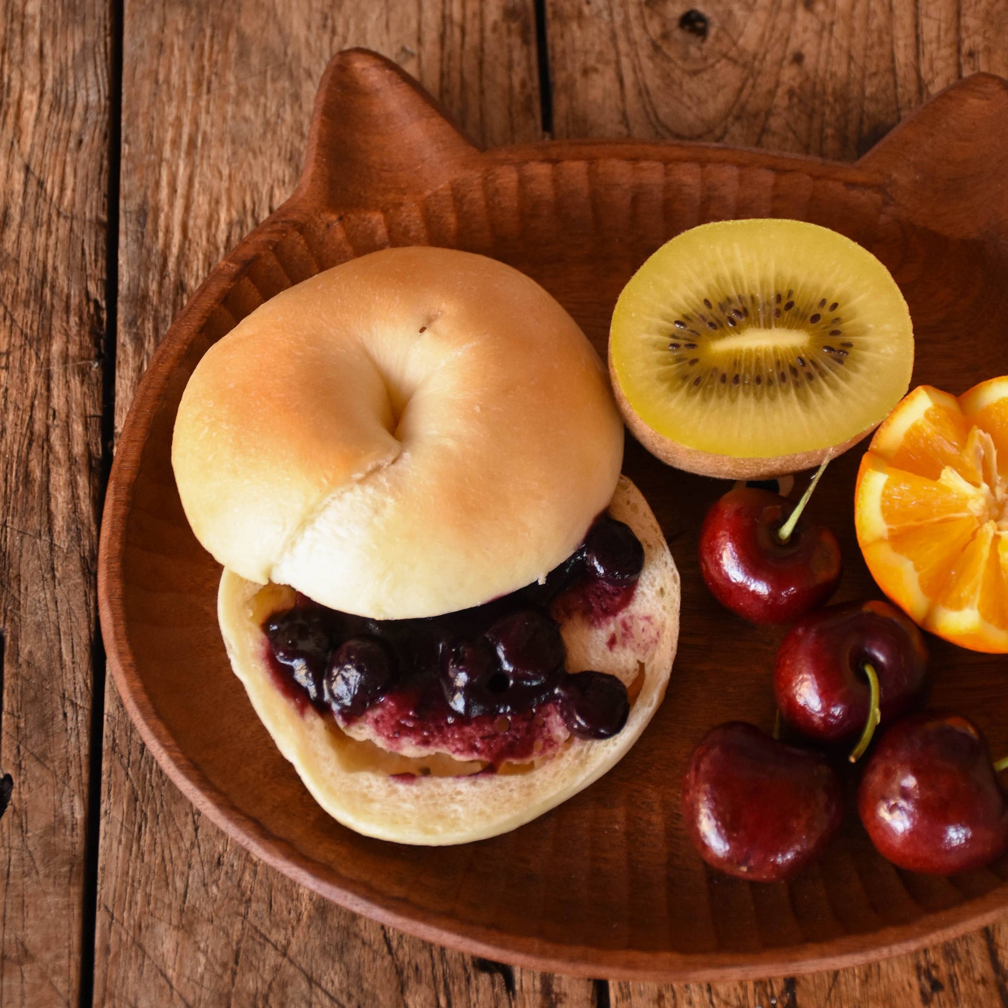 Bagel with jelly, kiwi, cherries, and an orange on a wooden plate.