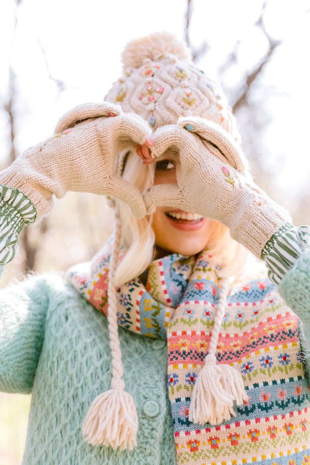 A smiling woman outdoors wearing a knitted beanie, cream knit gloves, and a multicolored Fair Isle scarf, holding her hands together in front of her face to form a heart shape.