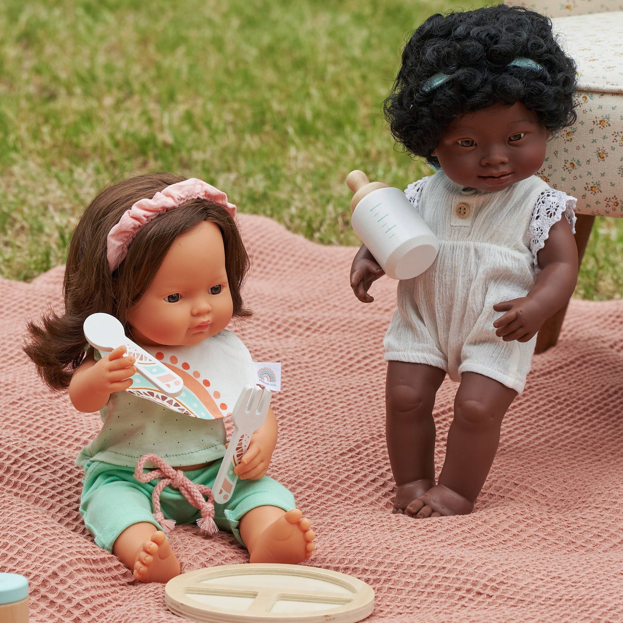 Two dolls, one sitting and one standing, on a pink blanket with a grassy background.