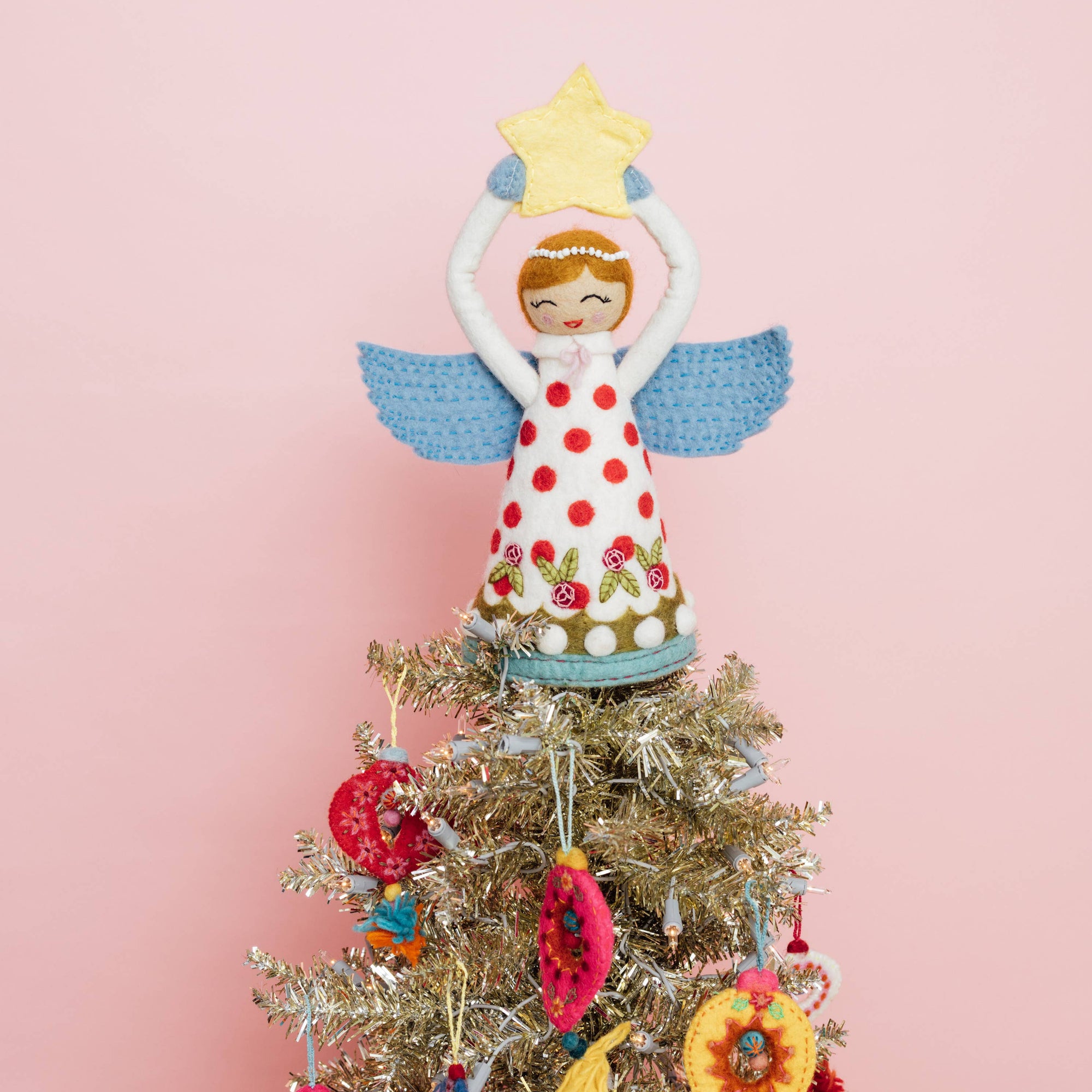 Decorative angel tree topper on a Christmas tree with colorful ornaments against a pink background
