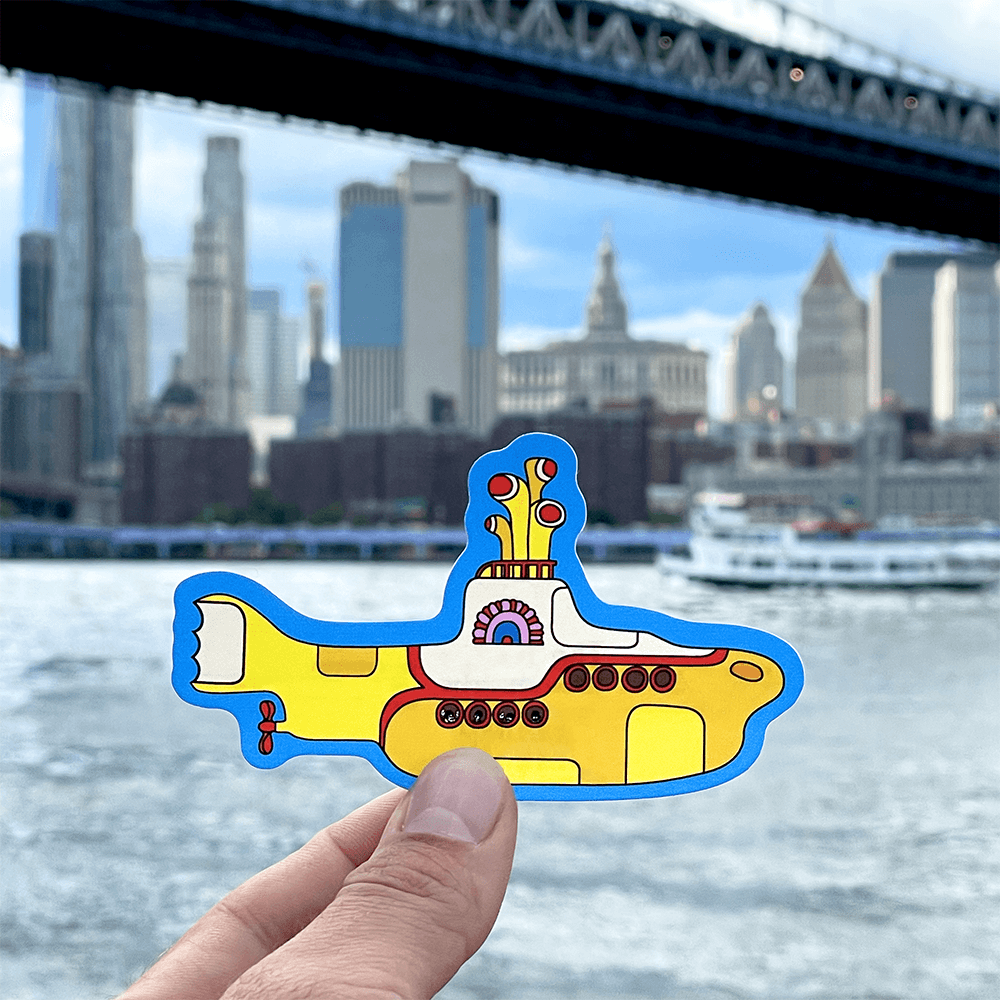 Colorful submarine sticker held up in front of a city skyline with a bridge and river.