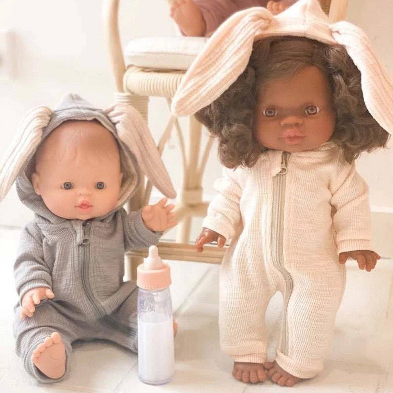 Two dolls in onesies with bunny ears, one holding a bottle, on a light wooden floor.